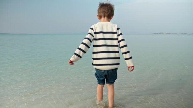 Little boy walking in the sea and looking on calm turquoise ocean waves. Holiday, vacation, weekend at nature