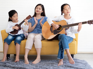 Pretty little girl sitting at couch enjoy playing acoustic guitar with her mother and sibling. Adorable kid playing ukulele with her sister and mom. Asian family guitarist singing together at leisure.