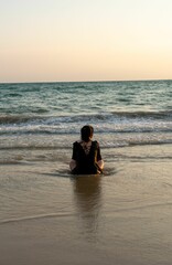 Woman sitting on a sandy shore of a beach, looking as the sun sets in the distance