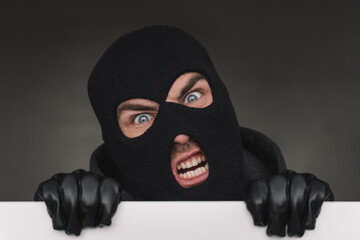An evil thief in a mask looks out from behind a white board. evil bandit with a mask on his face.