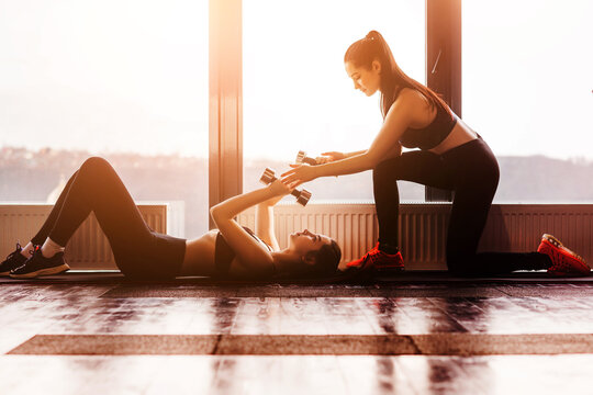 Two young female train together in modern gym on background of panoramic windows. Young teen girl is doing exercises with dumbbells lying on floor on back.
