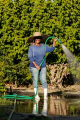 Vertical shot of a black woman wearing a hat and rain boots watering a garden