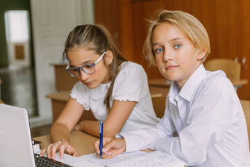 teen students working together writing in copybook using laptop in classroom