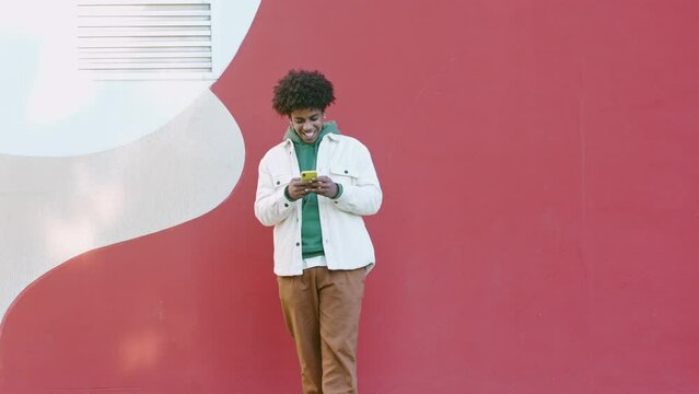 Cool young African American guy holding mobile phone standing at bright urban city wall. Happy stylish hipster teenager using cell outdoors, looking at cellphone texting message in social media chat.