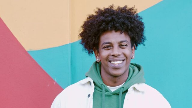Young happy African American teen guy laughing on colorful wall background. Smiling cool ethnic generation z teenager student model standing looking at camera posing for portrait outdoors.