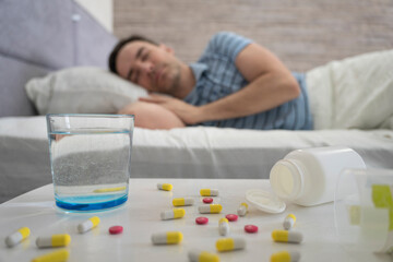Obraz na płótnie Canvas a man sleeps on a bed during the day in the foreground is a table with pills and a glass of water. Sleeping pills for a good sleep. Man sleeping - pills on bed table.