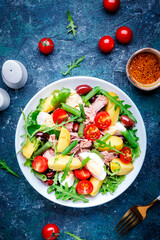 Nicoise salad with tuna, tomatoes, eggs, green beans, potatoes and olives on plate, blue table background, top view