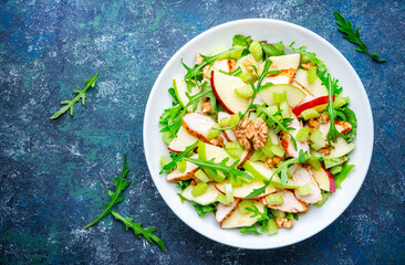 Waldorf salad with green and red apple, celery, lettuce, chicken fillet, arugula and crunchy walnuts on plate, blue table background, top view