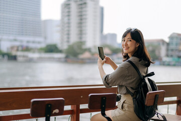 Fototapeta premium Young Asian woman backpack traveler using mobile phone in express boat pier on Chao Phraya River in Bangkok.