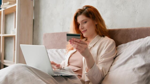 Happy female cardholder with credit card bank payment with laptop lying in bed at morning woman consumer buyer girl paying for purchase in web store online shopping computer ecommerce at home bedroom