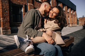 Portrait of beautiful emotional young couple in casual clothes smiling and hugging together while sitting on bench in city street