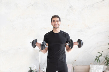 Smiling young handsome man in black sportswear doing biceps curl with dumbbells in living room at home. Healthy lifestyle and home workout concept