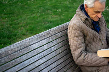 Old man with gray hair reading a book sited on an bench.