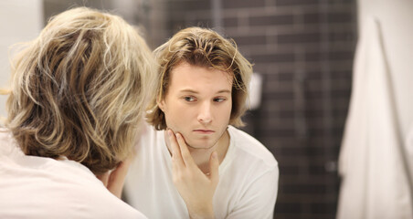 Fototapeta premium young man looking in the mirror,combing his hair,looking at problems on face.