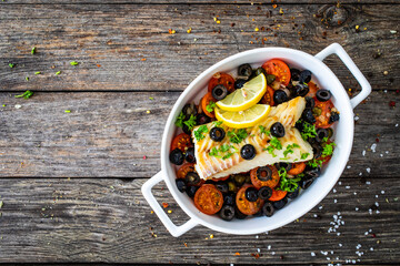 Cod in Italian style on tomatoes and black olives with baked potatoes in baking dish on wooden...