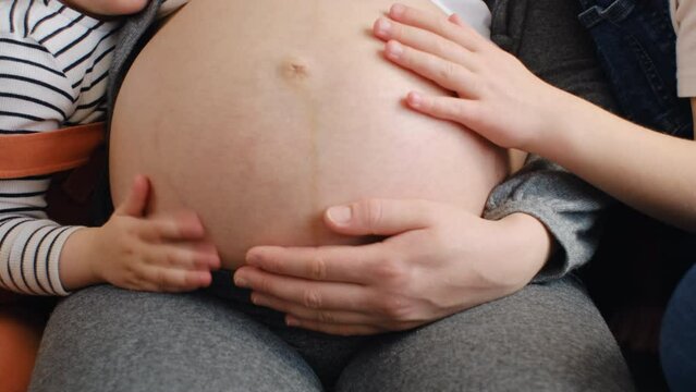 Close up of unrecognizable pregnant woman sitting on sofa with two little daughters, kids touches big belly of mother talking hugging enjoy tender moments and free time together. Motherhood concept