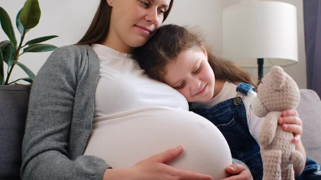 Positive engaged pregnant mom and smiling little daughter kid playing teddy bear toy on couch, interacting in role games, enjoying home activity, playtime, entertainment. Motherhood and family concept