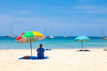 A lonely man sits under a colored umbrella in the shade from the hot sun on nai harn beach, thailand during vacation and travel. Beautiful scenery in Phuket.