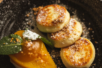 Syrniki with caramel pear and cottage cheese. Curd pancakes on black plate. Delicious breakfast. Ukrainian food. Baked pear with mint leaf and cheese pancakes. Restaurant menu. Homemade dessert.