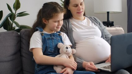 Caring young pregnant mother with cute little daughter sit on sofa using laptop spend weekend at home. Online content, educational program for kids, modern tech usage for purchasing or fun concept