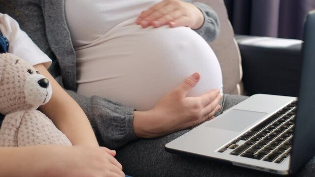 Close up of unrecognizable young pregnant mother with little daughter using laptop at home in living room. Mom watching cartoons on computer with small girl kid sitting on couch. Motherhood concept