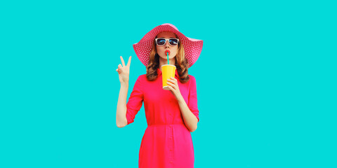 Summer portrait of beautiful young woman drinking fresh juice wearing straw round hat, pink dress on blue background