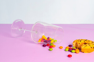 Fallen glass cup with a colorful round glazed candies and a bitten donut. isolated on pastel purple background.
