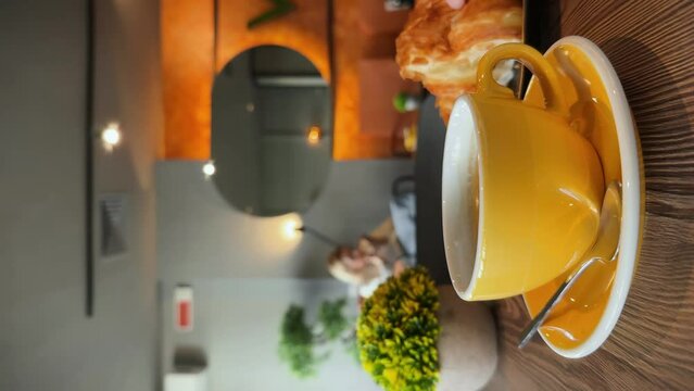 Eating fresh croissant with black coffee in a bakery. Dipping half of croissant into coffee, drops falling. People in bokeh, atmosphere of a coffee shop. Lifestyle vertical video.