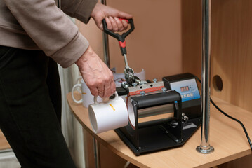 A woman at home learns to make souvenir cups on a heat press, a paper with a printout of an image for applying to a cup is attached to the cup with heat-resistant tape