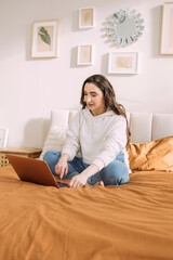 young curly girl in home casual clothes studying online at home sitting on the bed in her bedroom