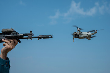 A man aims to shoot a rifle at a flying drone against a blue sky. 