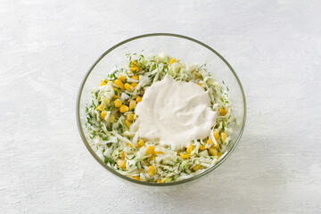 Glass bowl with salad of white cabbage, sweet corn, egg, dill and green onion with dressing on a light gray background, top view. Stage of cooking healthy vegan salad