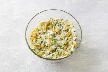 Glass bowl with salad of white cabbage, sweet corn, egg, dill and green onion with dressing on a light gray background, top view. Stage of cooking healthy vegan salad