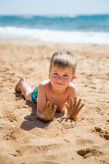 A boy in a swimming costume lies on his stomach on the sand by the sea and plays in the sand