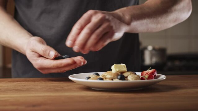 A man serving freshly made poffertjes with butter and fruits at home. Traditional Dutch mini pancakes with buckwheat flour, white flour, egg and milk.