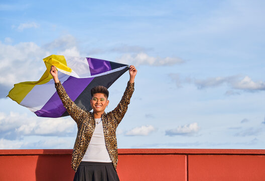 gender non binary asian person smiling holding gender non binary flag with blue sky background