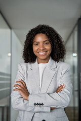 Happy smiling young African American leader manager, stand confident, crossed arms and looking at camera in business office center. Vertical portrait of professional business woman in stylish suit.