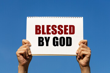 Blessed by god text on notebook paper held by 2 hands with isolated blue sky background. This message can be used as business concept about blessing.