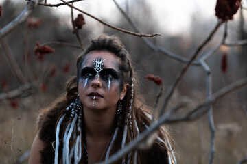 Autumn portrait of a female viking squatting in a forest. She is wearing face paint, leather and...