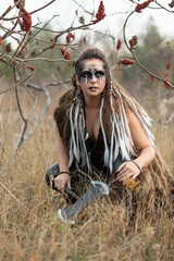 Autumn portrait of a female viking squatting in tall grass. She is wearing face paint, has her hair...