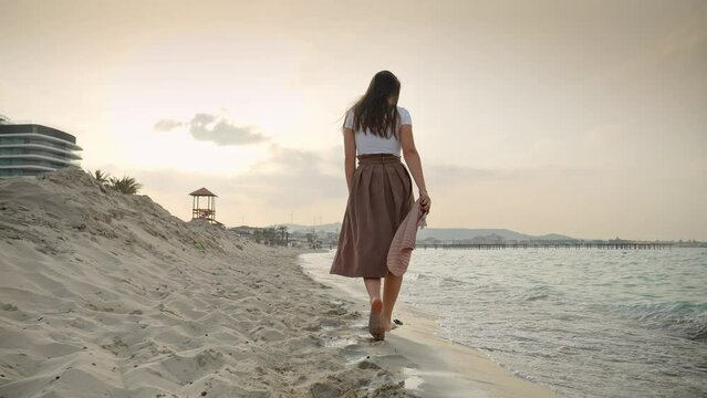 Slow motion of young woman with silk scarf walking on sandy sea beach at windy day towards the sunset. Concept of happiness, travel, journey, trip, tourism and holiday