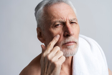 Fototapeta premium Serious senior man looking at mirror, after shower, touching face isolated on gray background