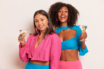 Fototapeta na wymiar Two young ladies having fun inside studio, black girl in blue top and Asian girl in pink stylish jacket, both girls hold glasses of cocktails, happy shopping time concept, copy space, high quality
