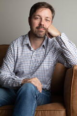 Portrait of a caucasian man sitting on a couch. He is in his 40s and is wearing a button up checked shirt. He is looking to his side. He is in the living room. 