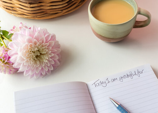 High angle closeup of notebook page with "Today I am grateful for" written on it, next to pink dahlia and cup of tea (selective focus)