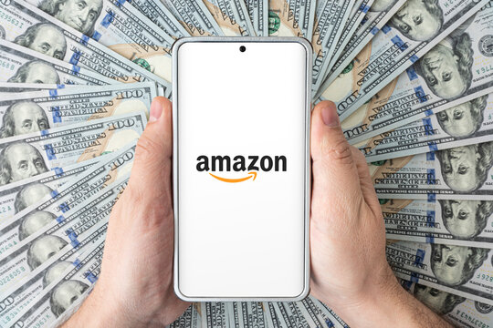 the logo of the amazon website on the smartphone screen in men's hands lying on American dollars.