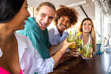 Hispanic young woman and multiracial friends toasting with mojito cocktails at a beach bar.