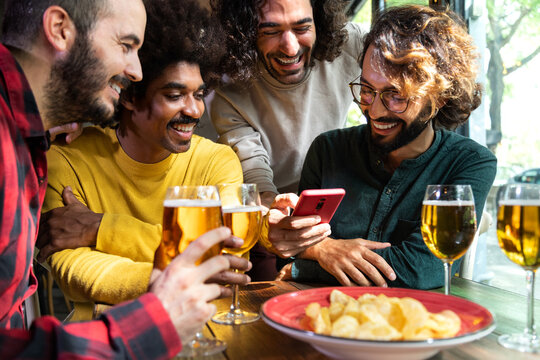 Multiracial guy friends looking at mobile phone together while having drinks together in pub. Vertical image