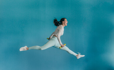 Young brunette American woman in sportswear jumps high holds dumbbells screams against blue wall....
