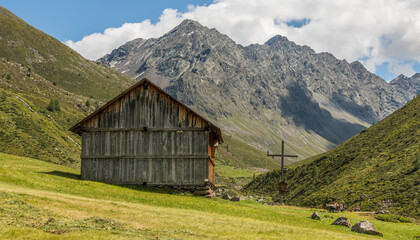 Fototapeta na wymiar Summer alpine view of a wooden barn in a fresh alpine meadow barn and a wooden cross. Mountains in the background. Umhausen, Austria